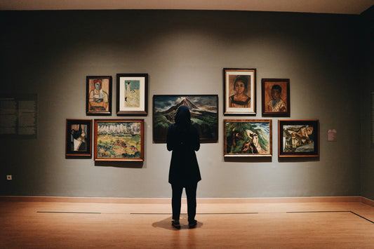 Person standing alone in a gallery. A spotlight focuses on them alone. A wall of framed paintings are hanging in front of them. 